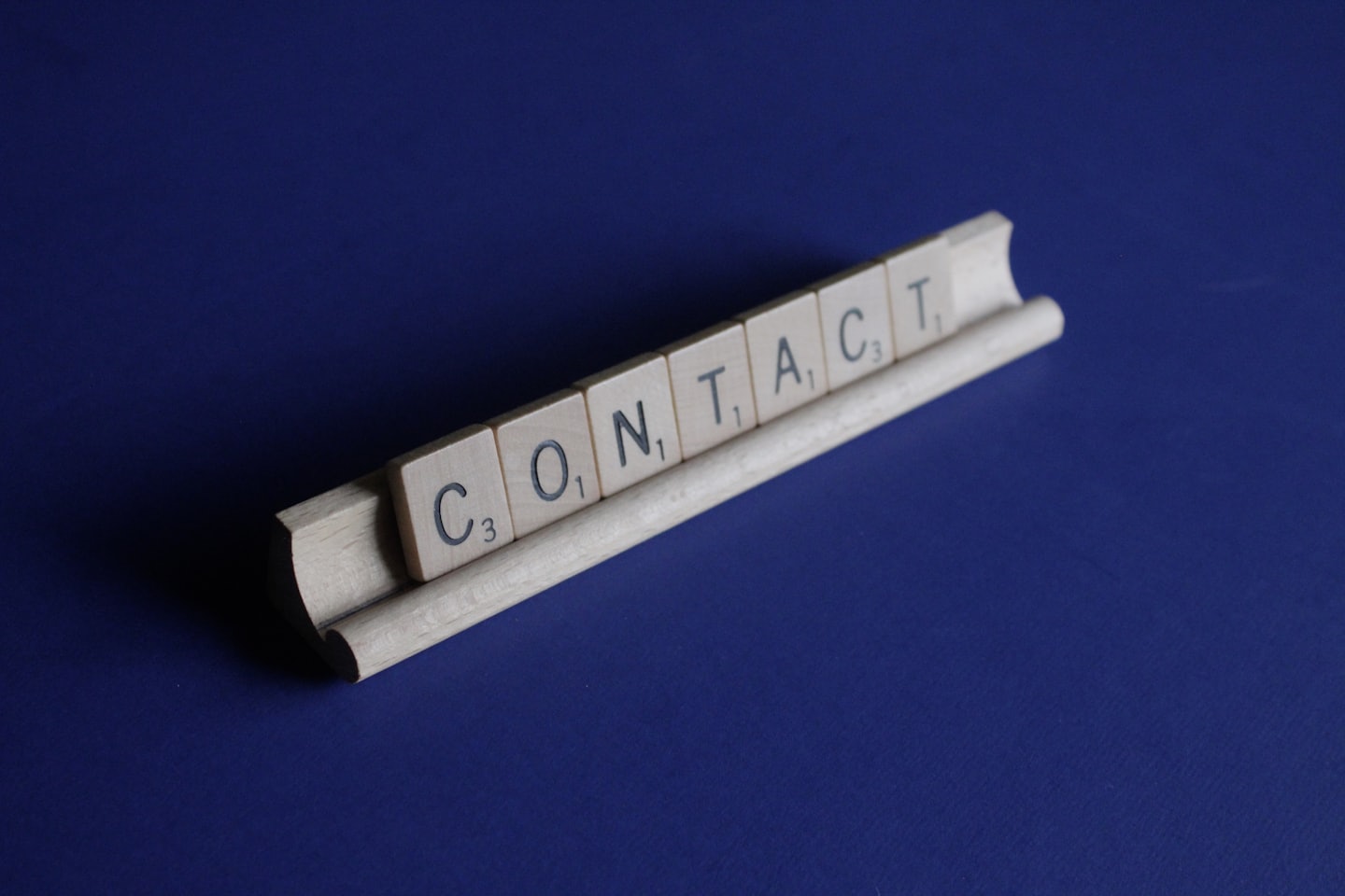 Scrabble tiles spell 'CONTACT' laid along a curved white wooden rod on a deep blue background.
