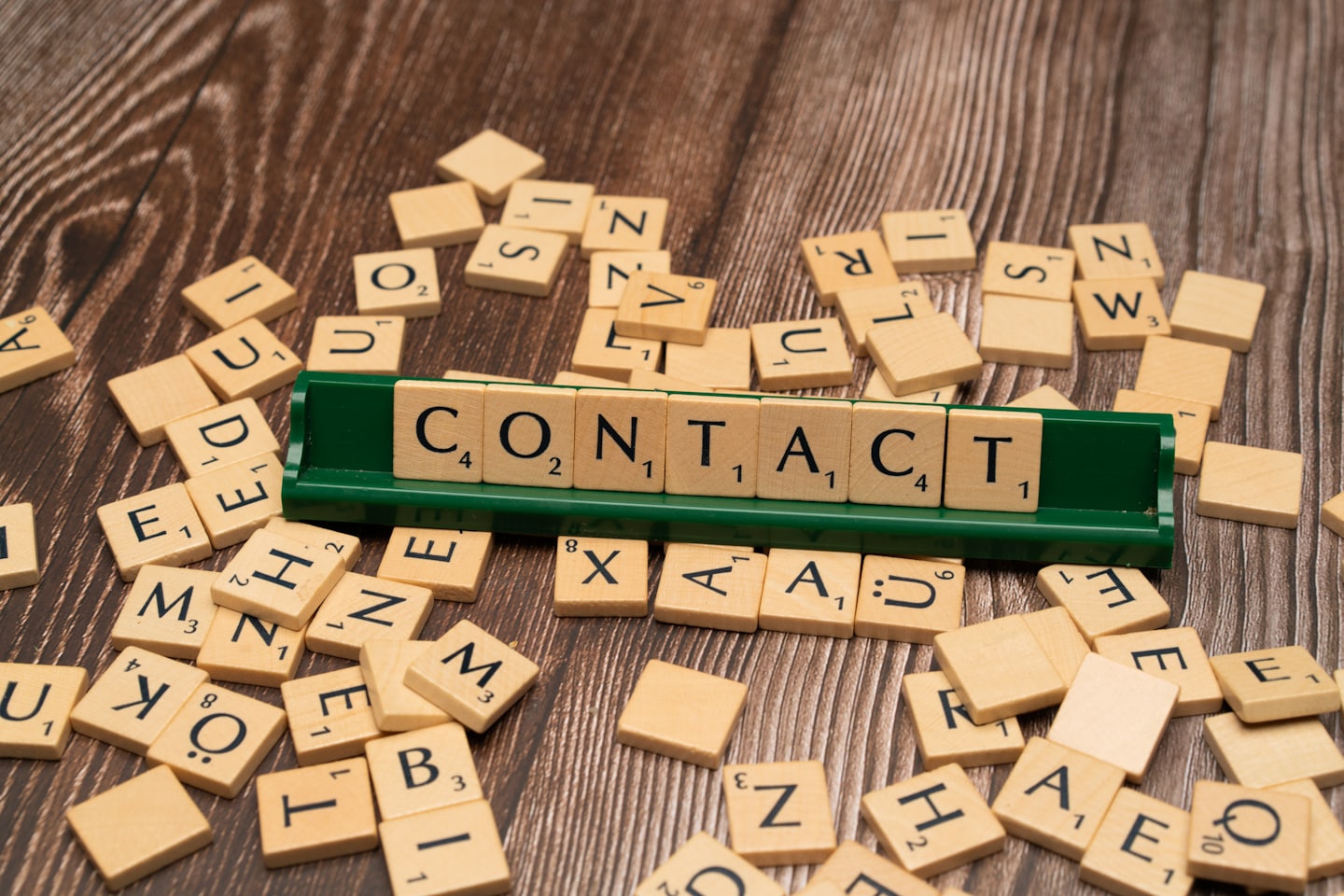 Scrabble tiles on a wooden table spell the word CONTACT on a green tile rack, with many letters scattered around.