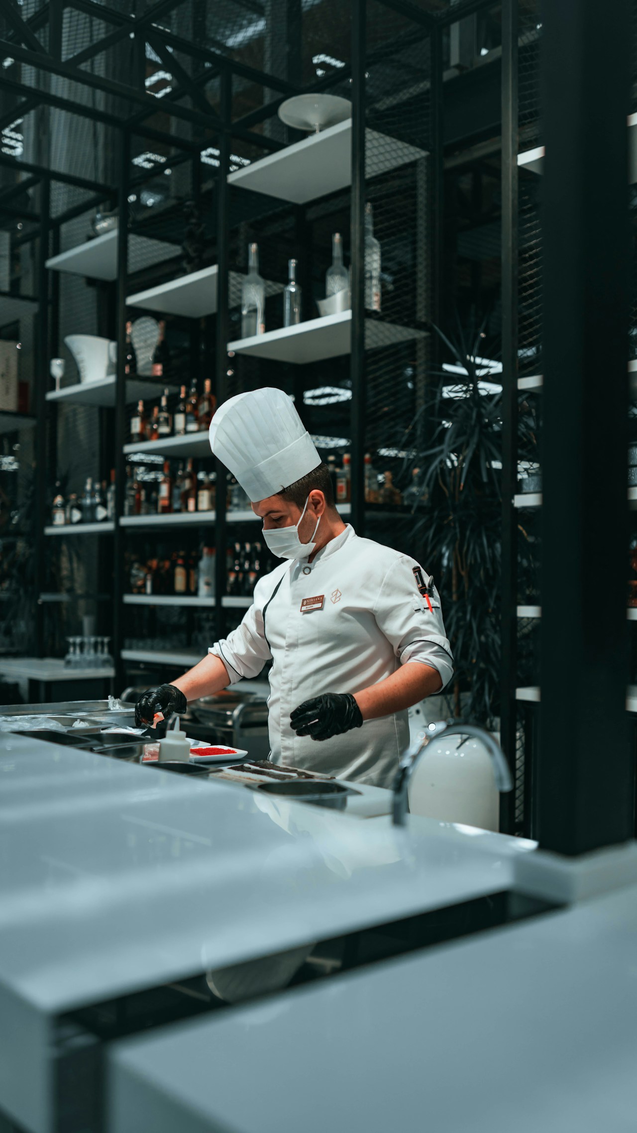 photo-1641536618422-2cf0bbd10014 - ChefHeads-Club der Küchenchefs Chef in a white uniform, tall hat and mask plating food at a stainless counter in a modern kitchen with bottles on shelves behind him.
