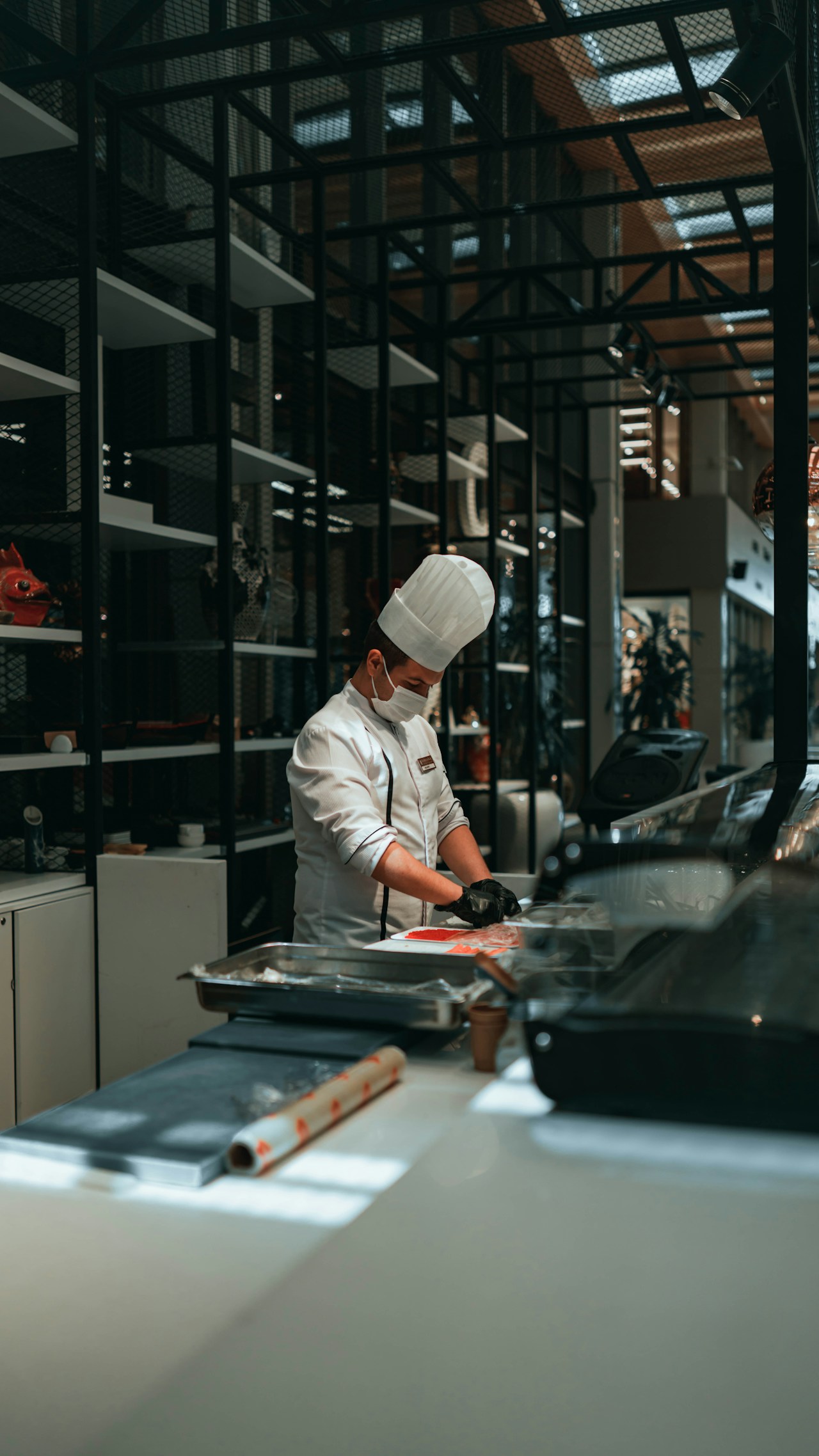 photo-1641536618779-b10c232ec2b2 - ChefHeads-Club der Küchenchefs Chef in white uniform and hat, wearing a mask, slicing fish at a seafood counter.