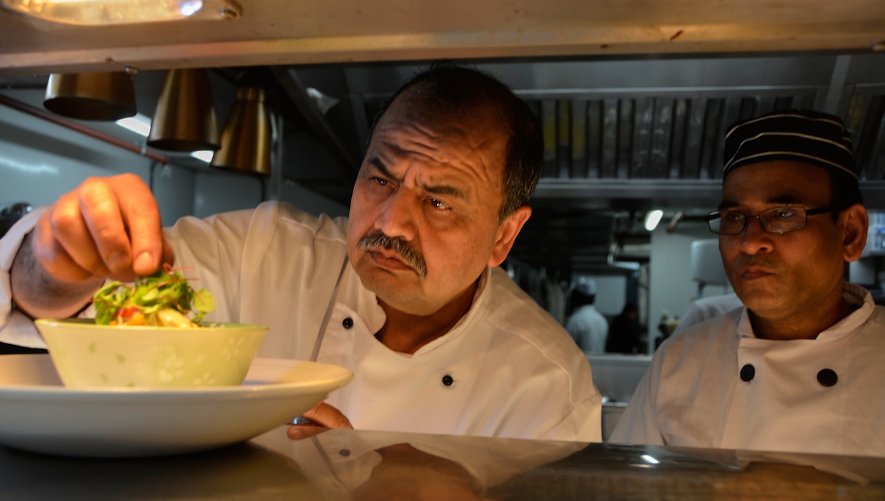 photo-1671029770042-201f223fba0f - ChefHeads-Club der Küchenchefs Head-and-shoulders view of a chef in a white uniform plating a salad, with another chef observing in a busy kitchen.