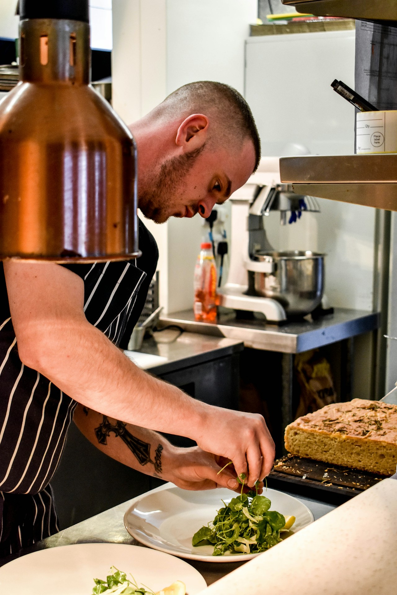 photo-1700484932777-b30323688070 - ChefHeads-Club der Küchenchefs Chef in a striped apron plating fresh greens on white plates in a busy kitchen set-up with a loaf of bread nearby