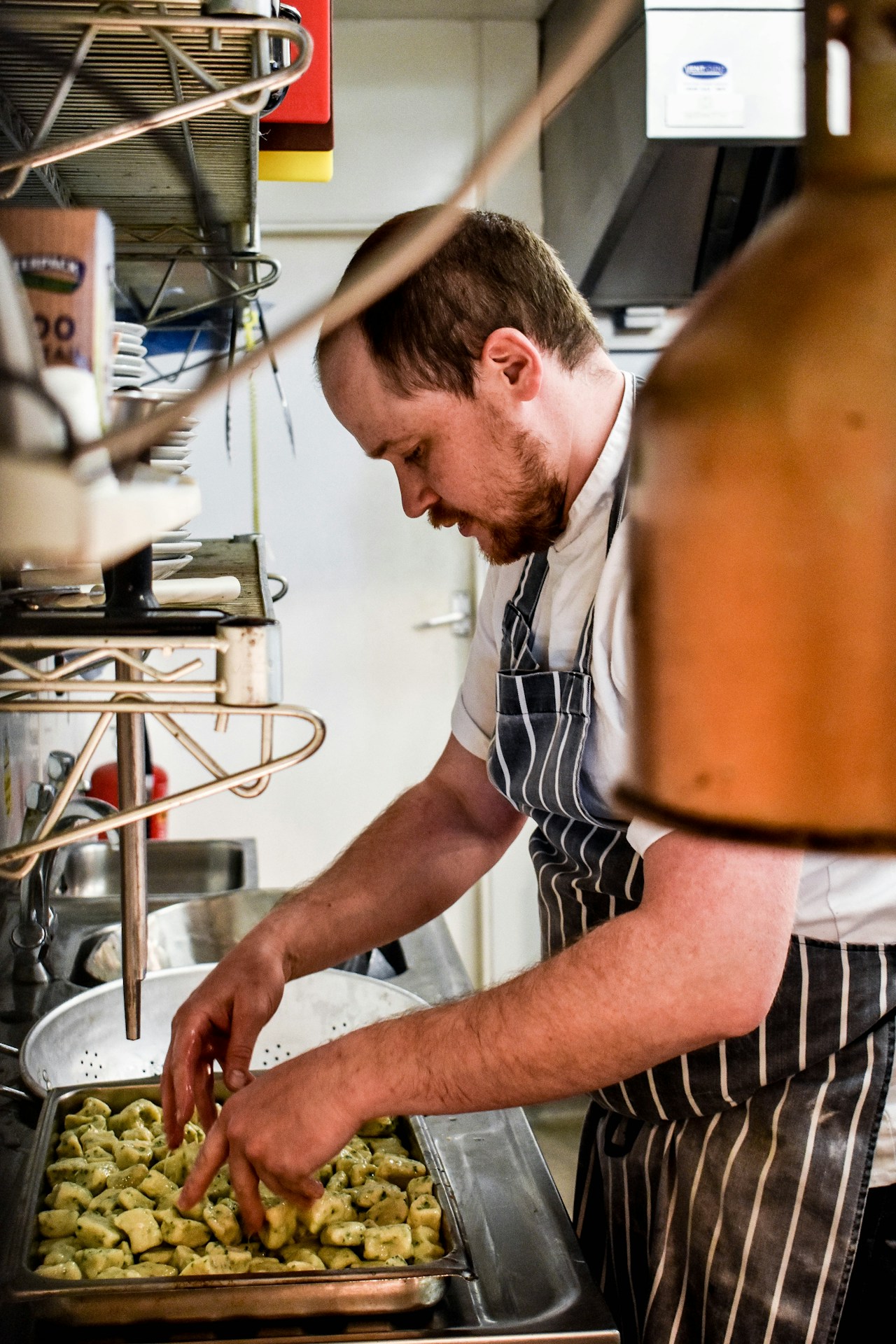photo-1700484932784-d083203b19be - ChefHeads-Club der Küchenchefs Chef in a striped apron shapes small dough dumplings onto a metal tray in a busy kitchen moment: focus on hands and tray.