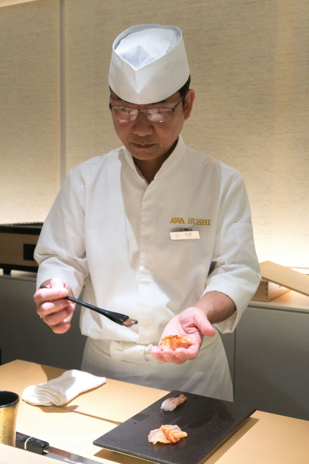 photo-1730635250911-b787fbc7e90f - ChefHeads-Club der Küchenchefs Sushi chef in white uniform and hat preparing nigiri at a counter, holding a piece of fish over rice on a black board.