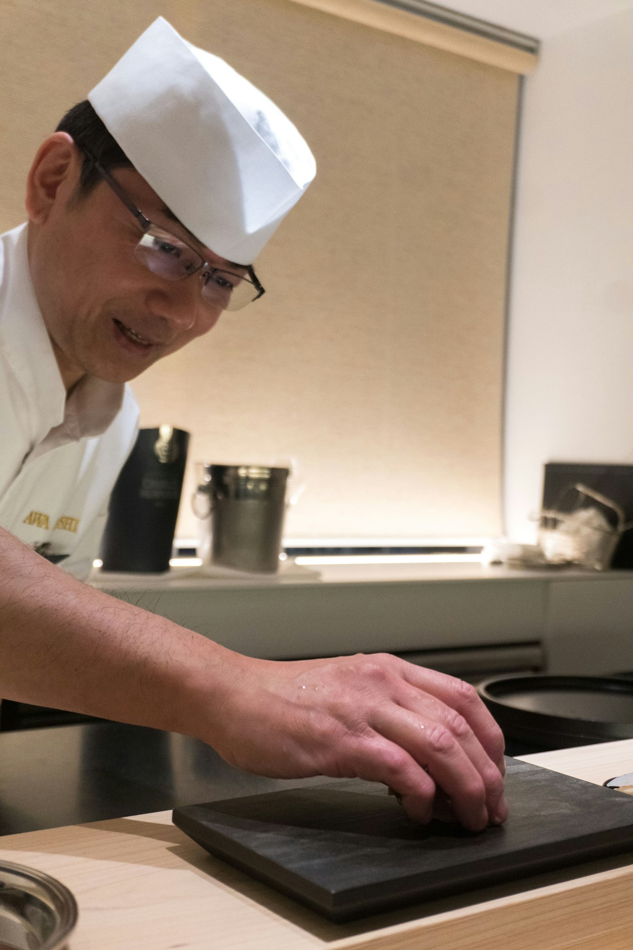 photo-1730635250971-c1ec966b0b11 - ChefHeads-Club der Küchenchefs Chef in white hat and jacket leaning over a kitchen counter, hands on a black slate board as he prepares food
