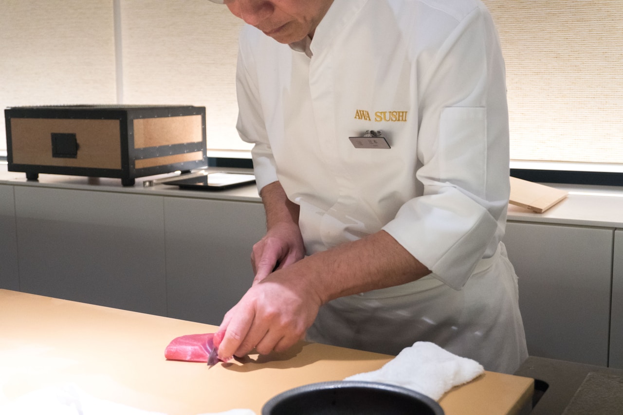 photo-1730635250973-aa696304add6 - ChefHeads-Club der Küchenchefs Sushi chef in a white uniform slices red tuna on a counter in a restaurant kitchen, focus on hands and knife.
