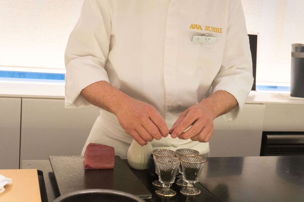 photo-1730635252093-2c586d3fefa0 - ChefHeads-Club der Küchenchefs Sushi chef in a white uniform shapes nigiri, with a slab of tuna on the counter and small glass cups nearby