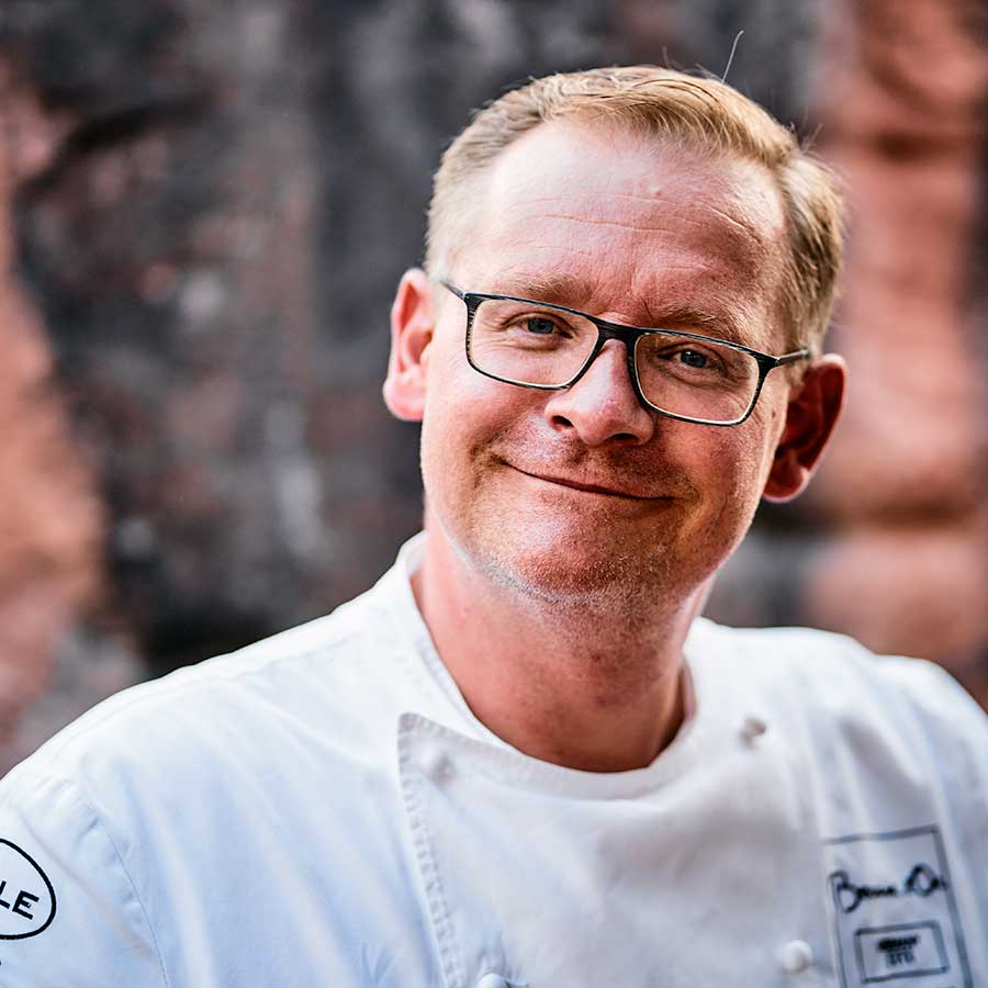 Smiling male chef wearing glasses and a white chef's coat, posing outdoors with a brick wall background.