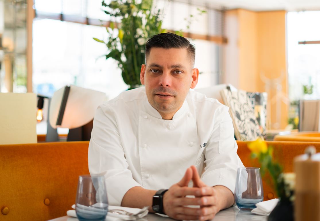 Chef in a white jacket sits at a table in a bright restaurant, hands folded, looking at the camera.