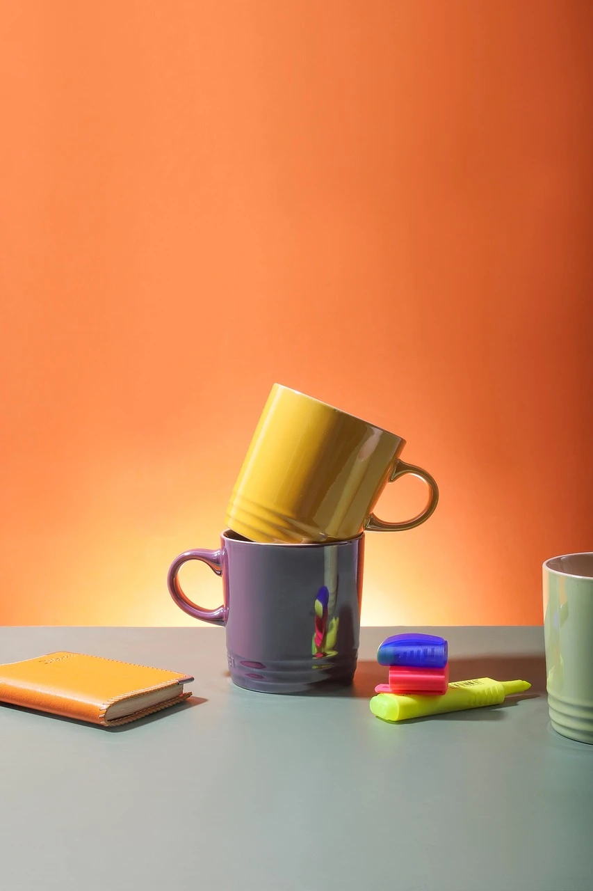 A yellow mug perched on a purple mug on a gray desk, with a notebook and bright highlighters nearby against an orange wall.