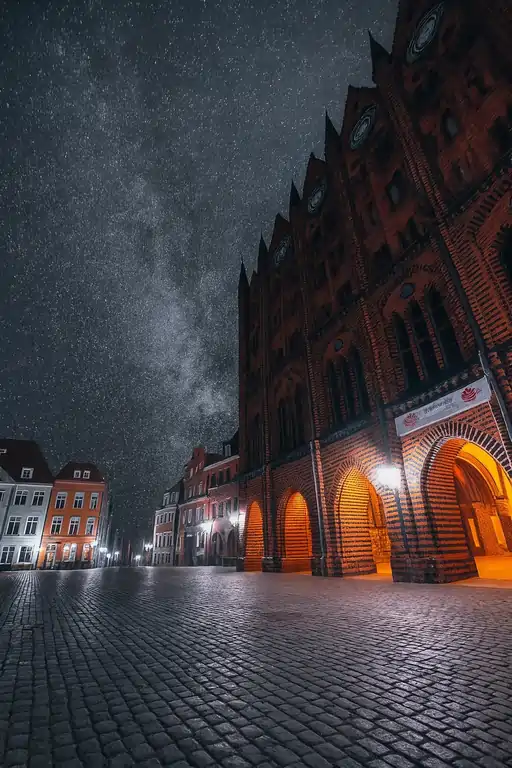 Night scene of a red brick Gothic building with arched doorways, glowing streetlights, and a starry sky over a wet cobblestone plaza.